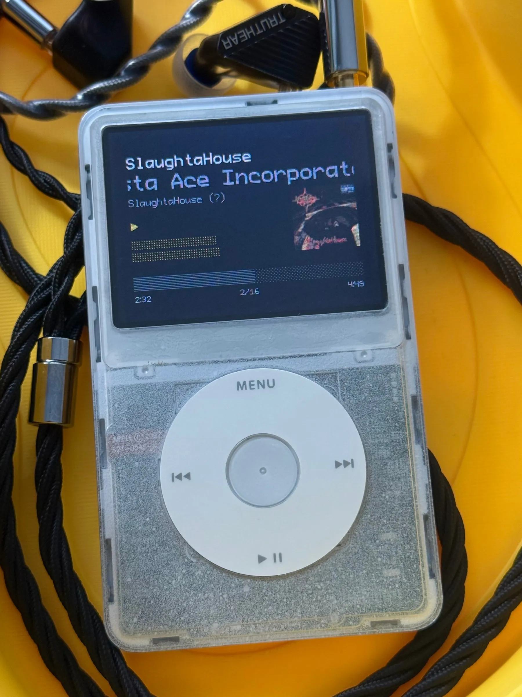 The iPod lying face up on a yellow surface, its clear shell and white click wheel ringed by a black braided cable and a pair of Truthear in-ear monitors, the screen playing SlaughtaHouse by Masta Ace Incorporated