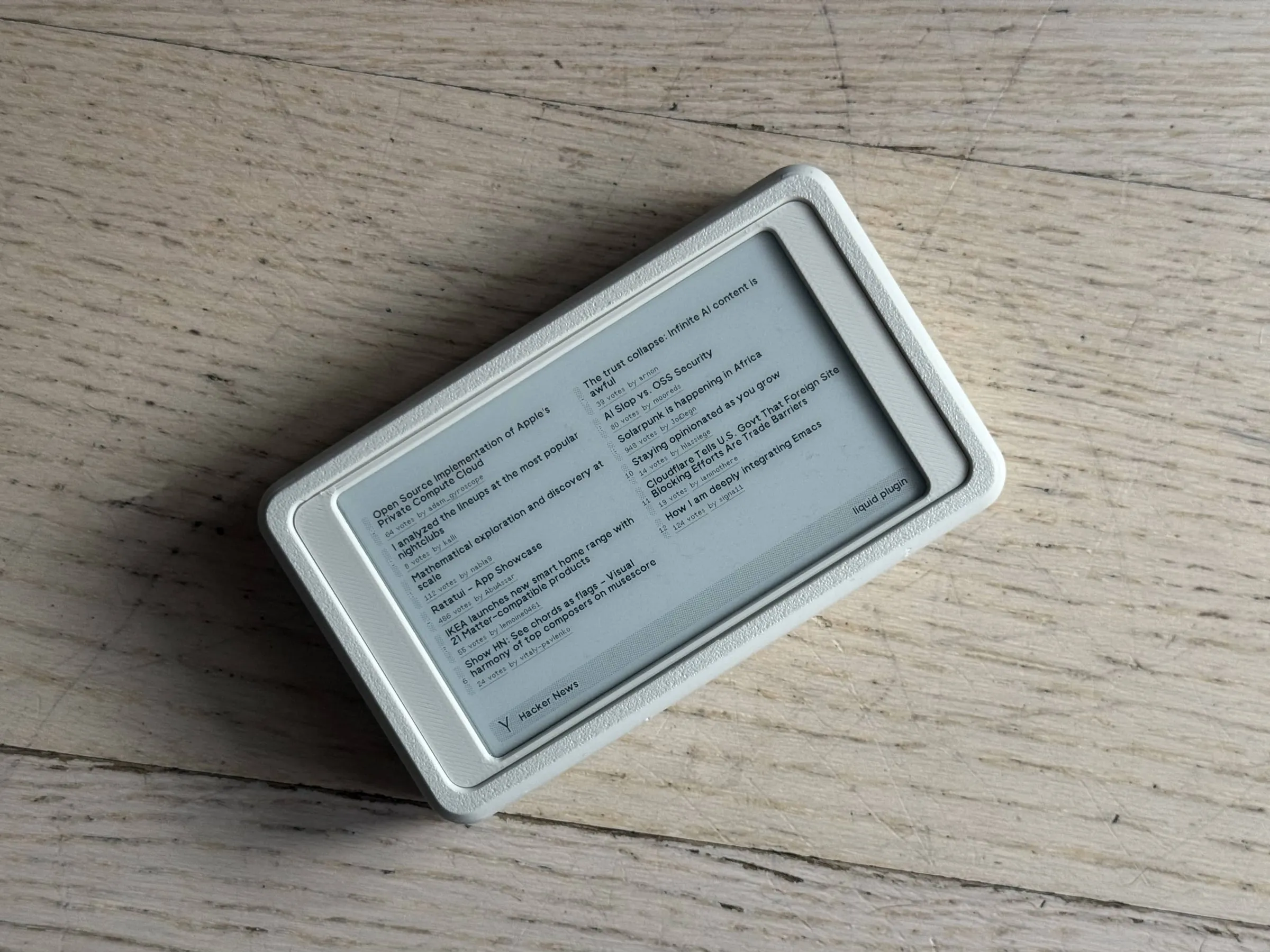 Small white e-ink display lying flat on a wooden floor, the screen showing Hacker News headlines in two columns