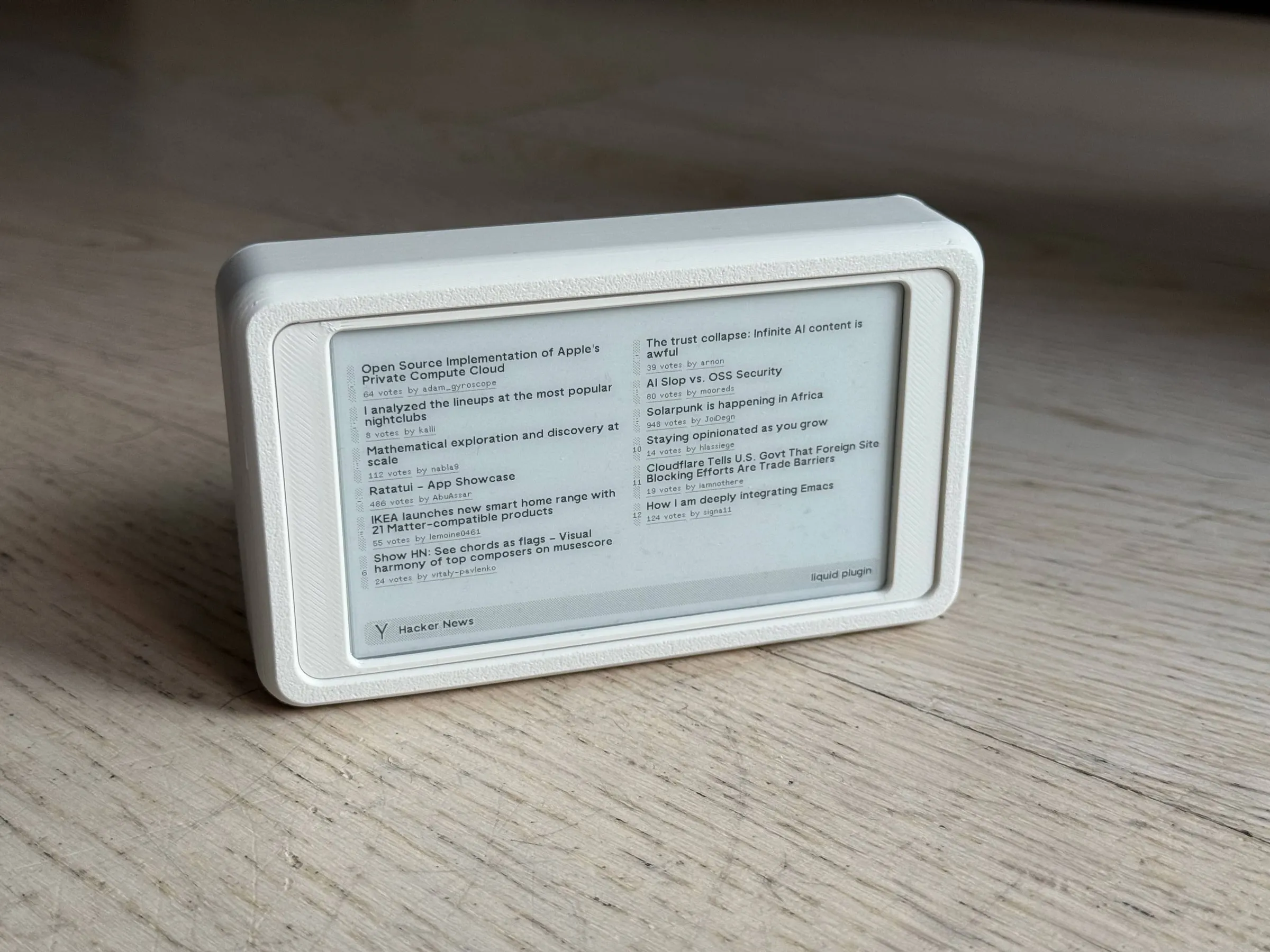 The mini standing on a wooden floor, seen from the front at an angle, the screen showing Hacker News headlines