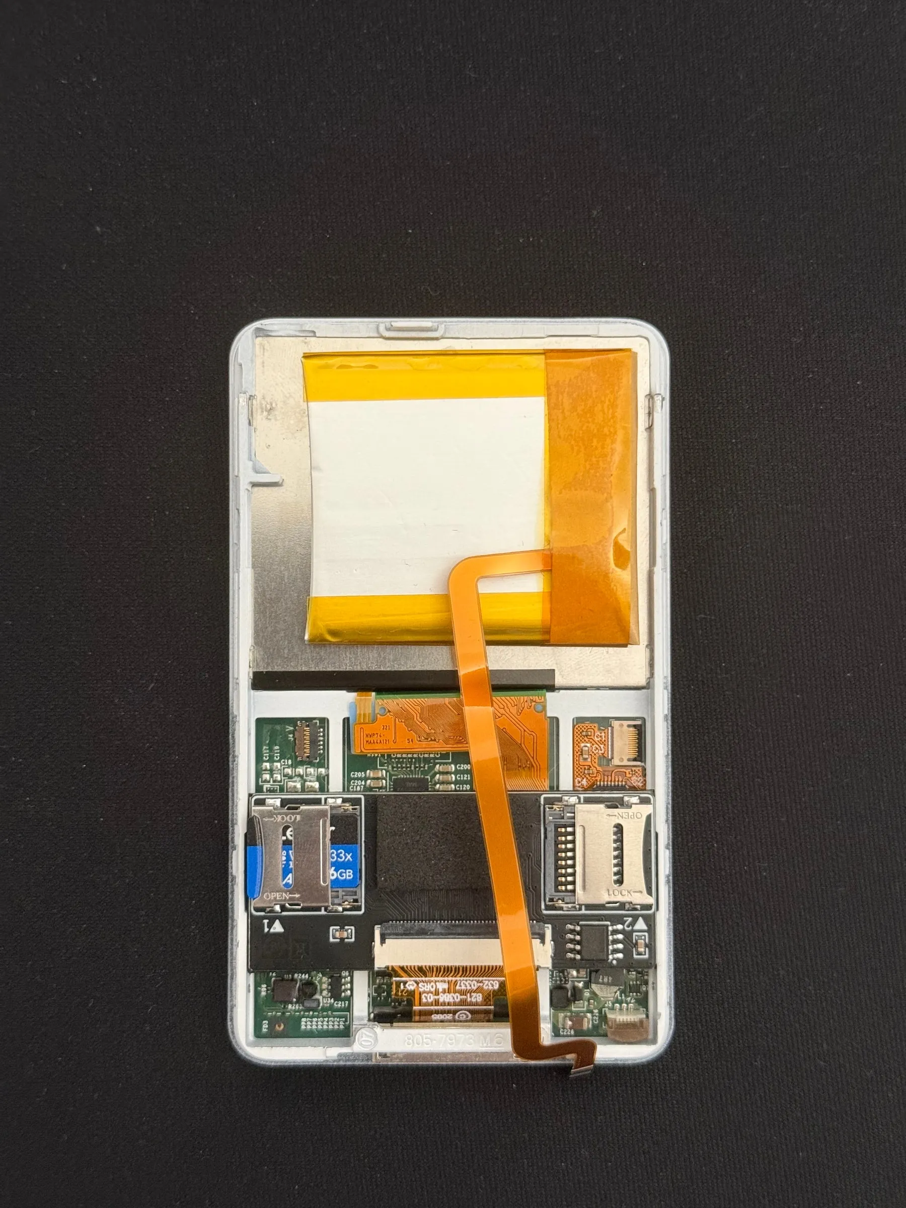 The rear half on its own, closer: the battery wrapped in white and yellow tape at the top, the logic board under it, the SD card in its adapter on the left and an orange ribbon cable running down the middle