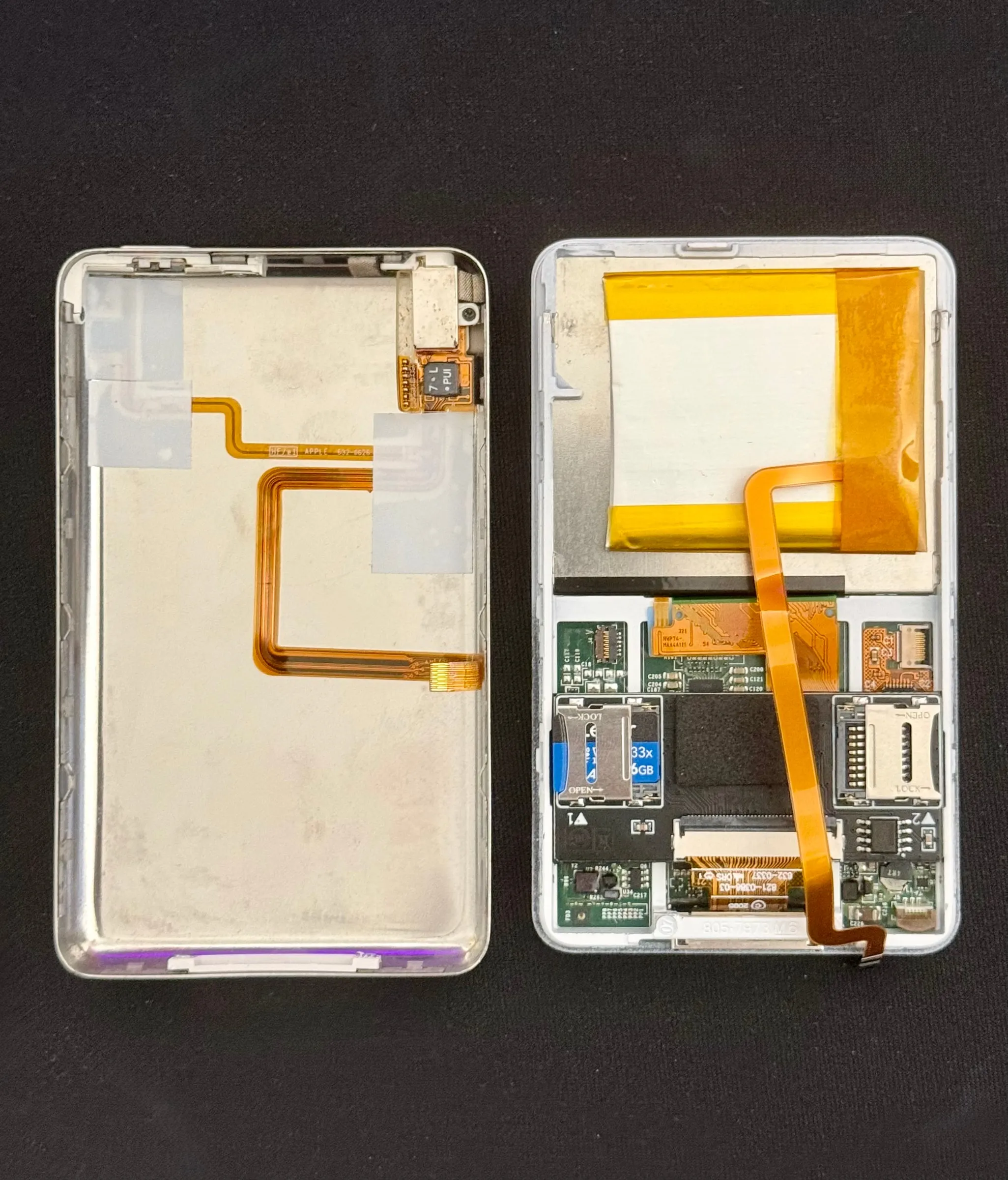 The iPod opened into its two halves on black fabric: on the left the front shell with the back of the screen and its orange ribbon cable, on the right the rear half with the battery taped in at the top, the logic board below it and an SD card in its adapter