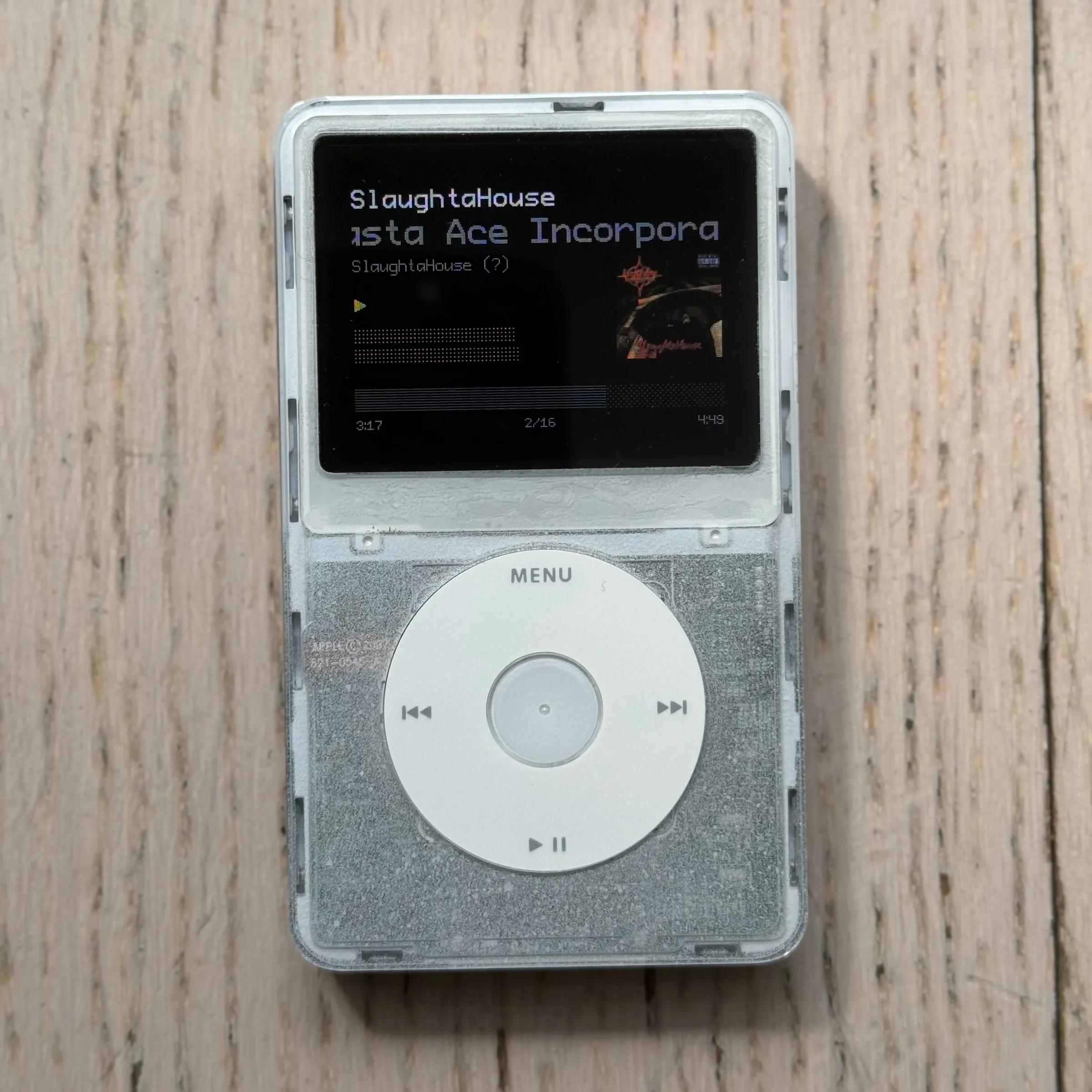 The finished iPod from the front on pale wood: a clear shell over a frosted white front, a white click wheel, and the now-playing screen showing SlaughtaHouse by Masta Ace Incorporated with album art, 3:17 of 4:49 played