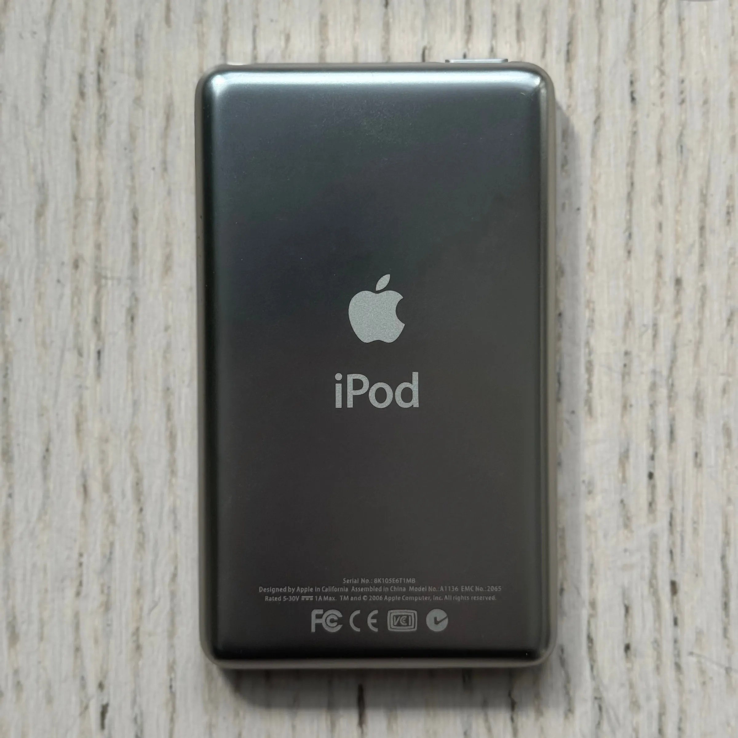 The back of the iPod on pale wood: a dark grey panel with a soft, non-mirror sheen, the Apple logo and the iPod wordmark in the middle and small print along the bottom