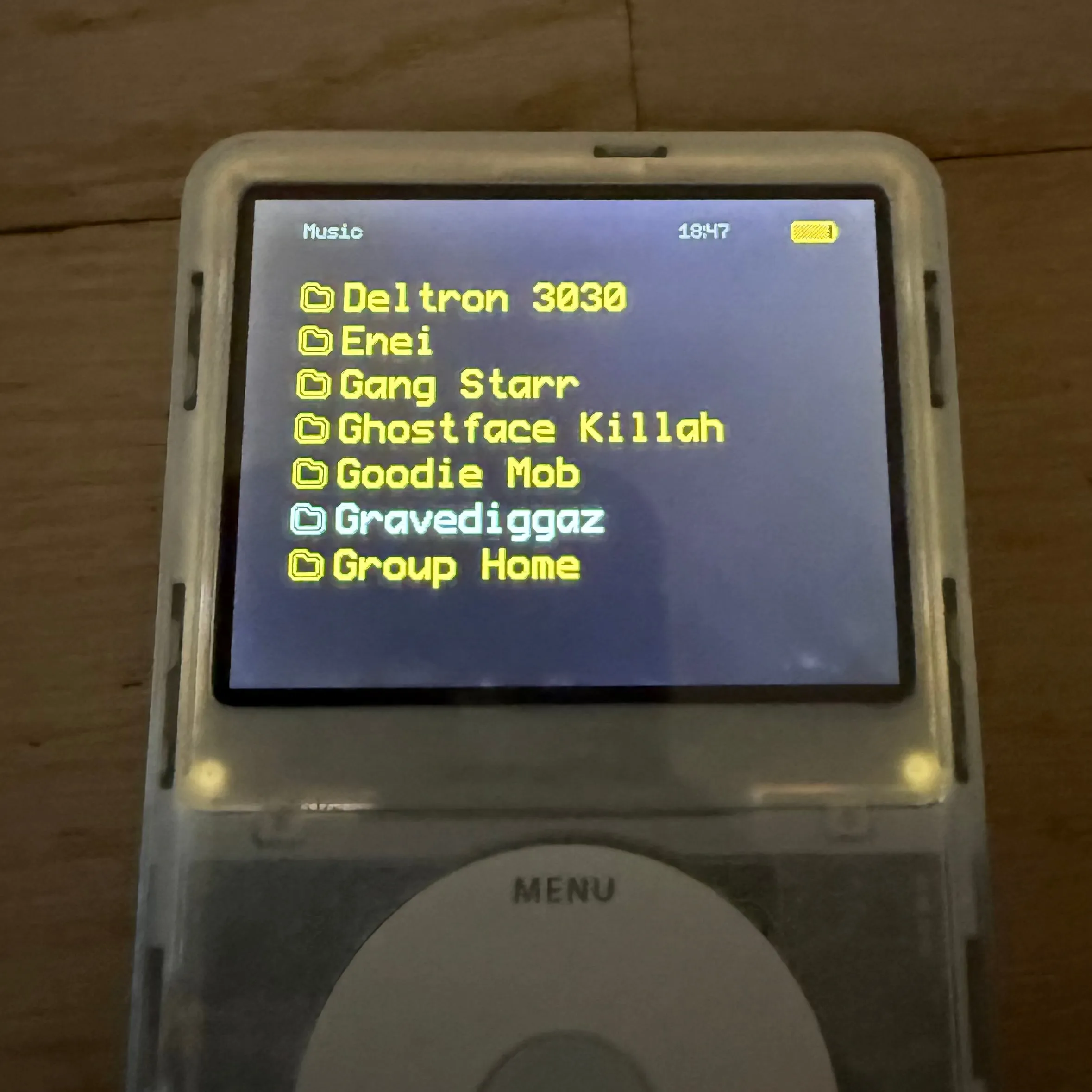 The iPod in a dim room showing the Music list in yellow on blue - Deltron 3030, Enei, Gang Starr, Ghostface Killah, Goodie Mob, Gravediggaz highlighted, Group Home - with two bright points glowing at the bottom corners of the screen where the backlight bleeds past it